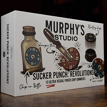 A boxed product labeled 'Murphy's Studio Sucker Punch: Revolutions' sits on a wooden surface in a dim indoor setting with a warm light in the background. The box is white with illustrated graphics and text, and the product appears to be a novelty or themed boxed item.