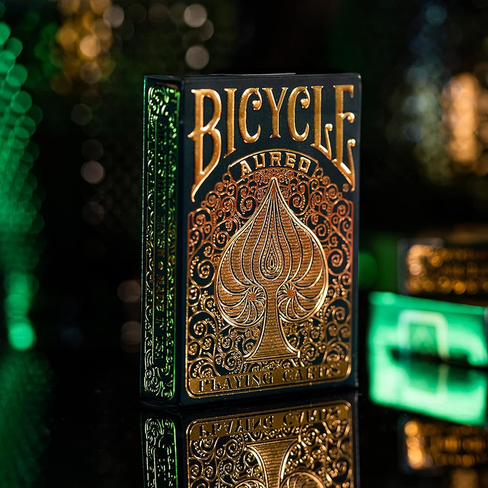 A pack of Bicycle Aureo Gold Playing Cards with gold foil and swirl designs on the tuck box, placed on a reflective surface with a blurred background.