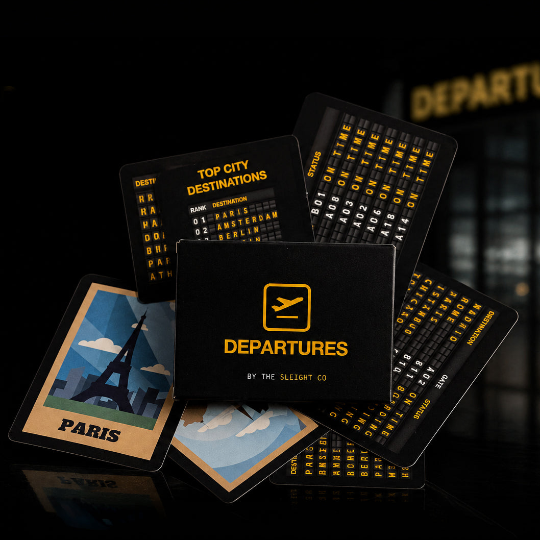 A set of black airport-themed cards and prints fanned out on a dark reflective surface, including a central card reading "DEPARTURES BY THE SLEIGHT CO," a Paris illustration with "PARIS," flight destination lists, and departure boards. The background is dark with a blurred airport-style sign.